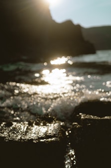 The photograph captures sunlight reflecting off ocean waves near a rocky shoreline. The background includes a silhouette of a cliff or rocky formation and the sun is partially visible at the top. The focus is soft, creating an ethereal, dreamy effect with shimmering light on the water's surface.
