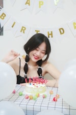 A young woman with braided hair is sitting at a table decorated for a birthday celebration. In front of her, there's a cake with colorful candies and pink candles spelling 'Happy Birthday'. Surrounding her are white balloons, and the wall is adorned with bunting featuring the letters 'Happy Birthday'. She is smiling and holding a lollipop.