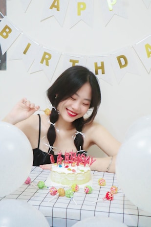 A young woman with braided hair is sitting at a table decorated for a birthday celebration. In front of her, there's a cake with colorful candies and pink candles spelling 'Happy Birthday'. Surrounding her are white balloons, and the wall is adorned with bunting featuring the letters 'Happy Birthday'. She is smiling and holding a lollipop.