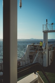 A person wearing a yellow safety vest stands on the deck of a ship, gazing at the serene water. The ship's railing and structures are visible, with distant mountains lining the horizon under a clear blue sky.
