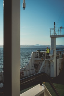 A person wearing a yellow safety vest stands on the deck of a ship, gazing at the serene water. The ship's railing and structures are visible, with distant mountains lining the horizon under a clear blue sky.
