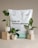 A white bag of soil labeled 'Erde für Zimmerpflanzen ohne Torf' is placed as the backdrop. In front, three potted green plants are displayed on beige pedestals of varying heights. The soil bag has branding and text in German and French, indicating that it is intended for indoor plants. The plants are in white, ribbed pots, and two are positioned on the pedestals for an aesthetically balanced arrangement.