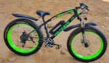A fat tire electric bike with a sleek black frame and vibrant green accents. The bike stands on sandy ground and features large, thick tires with 'XTRA ACTION' branding on the sidewalls. The seat is cushioned and ergonomic, designed for comfort.