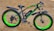 A fat tire electric bike with a sleek black frame and vibrant green accents. The bike stands on sandy ground and features large, thick tires with 'XTRA ACTION' branding on the sidewalls. The seat is cushioned and ergonomic, designed for comfort.