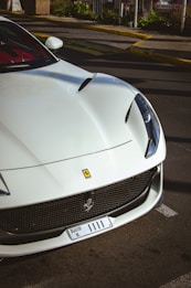 A luxurious white sports car is parked in an urban setting, with distinctive features including sleek lines and a prominent front grille. The car carries a Dubai license plate, and the background suggests a city environment with visible road markings and landscaping.