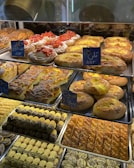 A display of various baked goods in a bakery. There are pastries topped with fresh strawberries, flaky danishes with custard, and assorted cookies with chocolate and pistachio. The shelves are filled with different types of breads and sweets, each labeled with small signs.
