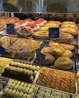 A display of various baked goods in a bakery. There are pastries topped with fresh strawberries, flaky danishes with custard, and assorted cookies with chocolate and pistachio. The shelves are filled with different types of breads and sweets, each labeled with small signs.