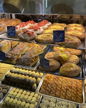 A display of various baked goods in a bakery. There are pastries topped with fresh strawberries, flaky danishes with custard, and assorted cookies with chocolate and pistachio. The shelves are filled with different types of breads and sweets, each labeled with small signs.