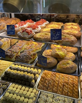 A display of various baked goods in a bakery. There are pastries topped with fresh strawberries, flaky danishes with custard, and assorted cookies with chocolate and pistachio. The shelves are filled with different types of breads and sweets, each labeled with small signs.