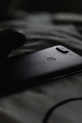 A smartphone with a dual camera setup and a fingerprint sensor on the back is lying on a soft, textured surface, likely a bed or blanket. The image is dark, with soft shadows around the phone, creating a moody atmosphere.