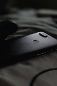 A smartphone with a dual camera setup and a fingerprint sensor on the back is lying on a soft, textured surface, likely a bed or blanket. The image is dark, with soft shadows around the phone, creating a moody atmosphere.
