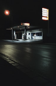 A dimly lit, empty gas station at night with a glowing streetlamp illuminating the wet pavement. The station features two gas pumps and a small building with a sign reading 'Walther'. There are tall signs displaying fuel prices, and the scene is engulfed in darkness except for the artificial lighting.
