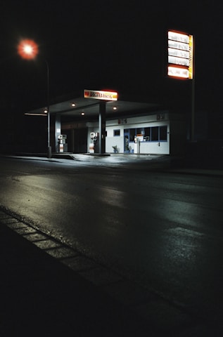 A dimly lit, empty gas station at night with a glowing streetlamp illuminating the wet pavement. The station features two gas pumps and a small building with a sign reading 'Walther'. There are tall signs displaying fuel prices, and the scene is engulfed in darkness except for the artificial lighting.