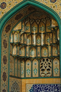 Intricate Islamic architectural details featuring ornate geometric patterns and calligraphy. The structure is adorned with arches and vibrant tile work, predominantly in teal, gold, and blue hues. The craftsmanship highlights traditional artistic elements in symmetrical designs.