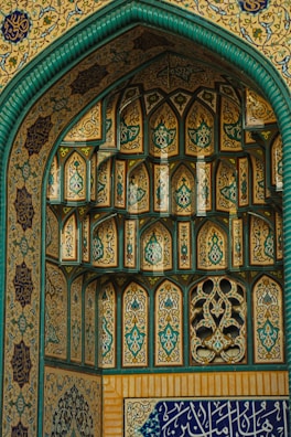 Intricate Islamic architectural details featuring ornate geometric patterns and calligraphy. The structure is adorned with arches and vibrant tile work, predominantly in teal, gold, and blue hues. The craftsmanship highlights traditional artistic elements in symmetrical designs.