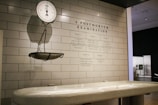 A clinical setting featuring an antique weighing scale suspended on a tiled wall. The scale is positioned above a white examination table. On the wall, there is detailed text titled 'A Postmortem Examination' outlining various procedural steps. The environment has a sterile and historical look, suggestive of a museum or educational display.
