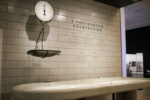 A clinical setting featuring an antique weighing scale suspended on a tiled wall. The scale is positioned above a white examination table. On the wall, there is detailed text titled 'A Postmortem Examination' outlining various procedural steps. The environment has a sterile and historical look, suggestive of a museum or educational display.