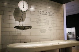 A clinical setting featuring an antique weighing scale suspended on a tiled wall. The scale is positioned above a white examination table. On the wall, there is detailed text titled 'A Postmortem Examination' outlining various procedural steps. The environment has a sterile and historical look, suggestive of a museum or educational display.