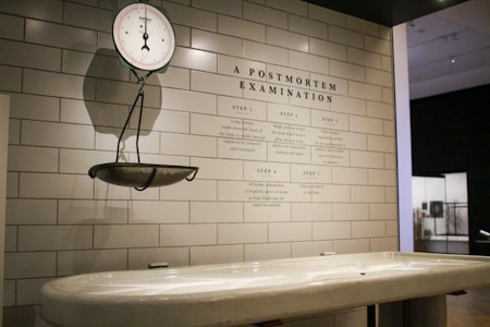 A clinical setting featuring an antique weighing scale suspended on a tiled wall. The scale is positioned above a white examination table. On the wall, there is detailed text titled 'A Postmortem Examination' outlining various procedural steps. The environment has a sterile and historical look, suggestive of a museum or educational display.