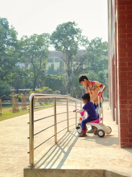 Two children are playing with a tricycle on a sunny day next to a building with a red brick wall. One child is sitting on the tricycle while the other child appears to be helping or guiding. In the background, there are green trees and a few buildings along with a metal railing beside the paved area.