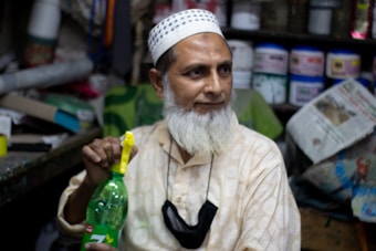 An older man with a white beard, wearing a patterned ivory kurta and a prayer cap, is seated indoors. He holds a green plastic bottle with a yellow handle in one hand. Shelves lined with various containers and bags are visible in the background, indicating a shop or cluttered workspace setting. The man looks content and relaxed.
