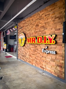 A storefront with a wooden panel wall featuring the bright yellow and red signage for MR.D.I.Y. Express. Flags are hanging from the ceiling inside the store, and a sale sign is visible near the entrance. The floor is concrete, and there's a sense of a clean, organized space.