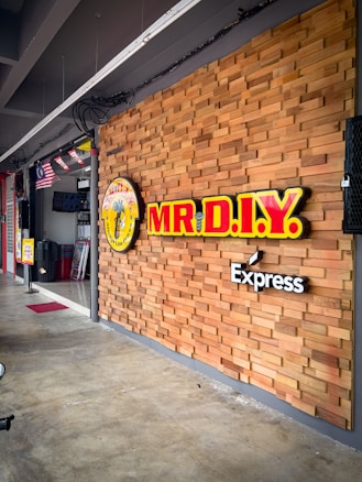 A storefront with a wooden panel wall featuring the bright yellow and red signage for MR.D.I.Y. Express. Flags are hanging from the ceiling inside the store, and a sale sign is visible near the entrance. The floor is concrete, and there's a sense of a clean, organized space.