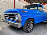 A vintage blue pickup truck is parked inside a garage-like space with a partially visible brick wall. The vehicle is well-preserved, showcasing a classic design with prominent features such as a chrome front grille and round headlights. The license plate is visible at the front. The truck's body is in good condition with shiny paint and retro side mirrors.