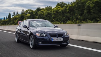 A sleek, black BMW car is driving on a highway surrounded by lush greenery and a cloudy sky. The road is wide and smooth, with a concrete barrier on the side. The car's headlights are on, and it has a European license plate.
