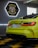 A vibrant green sports car is positioned inside a modern garage. The sleek design is highlighted by the bright red taillights. A sign reading 'TopCar Garage' is illuminated in the background with contrasting neon lights. The car is parked on a black and yellow checkered floor, enhancing the bold aesthetic.