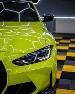A brightly colored neon green sports car with a sleek and modern design is parked indoors. The car's headlight is prominently displayed, featuring intricate LED details. The reflective paint surface mirrors the geometric ceiling lights. The floor consists of a black and yellow checkered pattern, adding to the boldness of the scene.