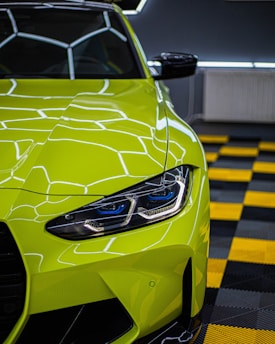 A brightly colored neon green sports car with a sleek and modern design is parked indoors. The car's headlight is prominently displayed, featuring intricate LED details. The reflective paint surface mirrors the geometric ceiling lights. The floor consists of a black and yellow checkered pattern, adding to the boldness of the scene.