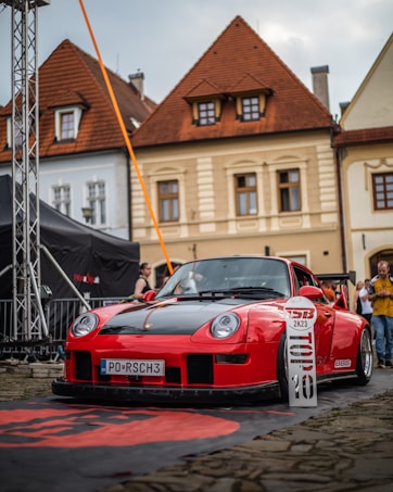 A red and black sports car is parked on a cobblestone street in front of a row of traditional European buildings with steep, gabled roofs. The car has a prominent front splitter and a license plate that reads 'PO RSCH3'. There are people in the background, and a banner on the ground in front of the car.