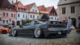 A modified sports car with an open hood and wide rims is parked on a cobblestone street. Several people are gathered around, with old European-style buildings in the background. The car has a sleek, black finish and a prominent rear spoiler.