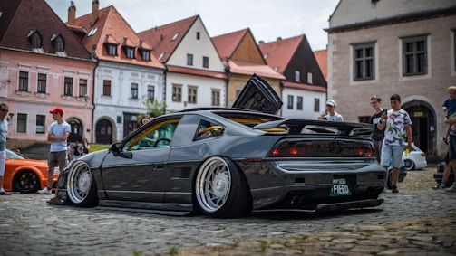 A modified sports car with an open hood and wide rims is parked on a cobblestone street. Several people are gathered around, with old European-style buildings in the background. The car has a sleek, black finish and a prominent rear spoiler.