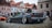 A modified sports car with an open hood and wide rims is parked on a cobblestone street. Several people are gathered around, with old European-style buildings in the background. The car has a sleek, black finish and a prominent rear spoiler.