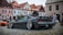 A modified sports car with an open hood and wide rims is parked on a cobblestone street. Several people are gathered around, with old European-style buildings in the background. The car has a sleek, black finish and a prominent rear spoiler.