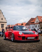 A red sports car with a black hood is parked on a cobblestone street in a European town. The car has a low profile, custom modifications, and BBS rims. In the background, people are walking by and medieval-style buildings with red-tiled roofs are visible under a partly cloudy sky.
