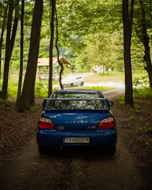 A blue Subaru car is parked on a dirt path in a forested area, surrounded by tall trees. In the background, another vehicle and a small building are visible along a road.