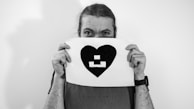 A person holding a piece of paper featuring a black heart with a geometric design inside it. The individual is partially obscured by the paper, which they hold up to their face, revealing only their eyes. They are wearing a casual shirt, and their long hair is tied back. The image is in black and white, creating a classic and timeless feel.
