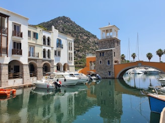 A picturesque marina with several boats docked along a calm waterway. The foreground shows a row of colorful buildings with arched windows and balconies. A stone tower with a pavilion top stands next to an orange arched bridge. In the background, palm trees line the waterway, and a verdant mountain rises under a clear blue sky.