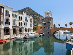 A picturesque marina with several boats docked along a calm waterway. The foreground shows a row of colorful buildings with arched windows and balconies. A stone tower with a pavilion top stands next to an orange arched bridge. In the background, palm trees line the waterway, and a verdant mountain rises under a clear blue sky.
