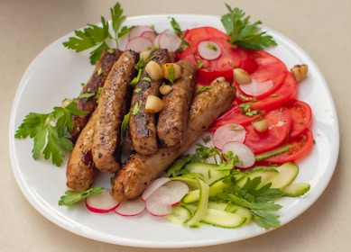A white plate filled with grilled sausages garnished with chopped herbs and garlic cloves. Slices of fresh tomatoes, radishes, and cucumber laid around the sides, with parsley leaves as additional garnish.