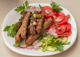 A white plate filled with grilled sausages garnished with chopped herbs and garlic cloves. Slices of fresh tomatoes, radishes, and cucumber laid around the sides, with parsley leaves as additional garnish.