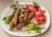 A white plate filled with grilled sausages garnished with chopped herbs and garlic cloves. Slices of fresh tomatoes, radishes, and cucumber laid around the sides, with parsley leaves as additional garnish.