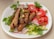 A white plate filled with grilled sausages garnished with chopped herbs and garlic cloves. Slices of fresh tomatoes, radishes, and cucumber laid around the sides, with parsley leaves as additional garnish.