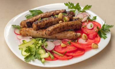 A white plate is filled with grilled sausages garnished with chives, surrounded by slices of fresh red tomatoes, radishes, and cucumber ribbons. Parsley leaves and small roasted garlic cloves add a touch of greenery and flavor. The presentation is both colorful and appetizing.