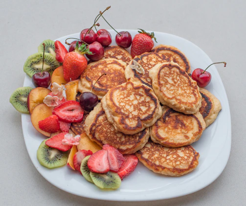 A colorful stack of millet pancakes topped with fresh fruits and nuts, surrounded by smiling children enjoying their snacks.