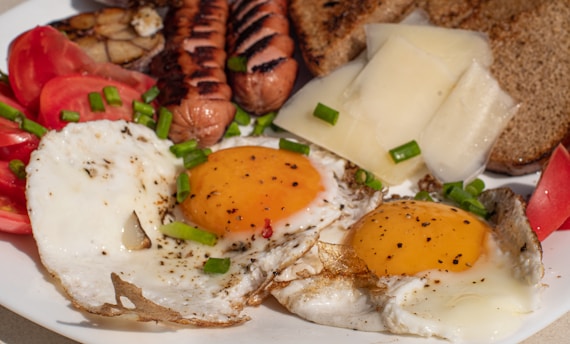 A breakfast plate consisting of two sunny-side-up eggs garnished with green onions, slices of tomatoes, grilled sausages, sliced cheese, and pieces of toasted bread.