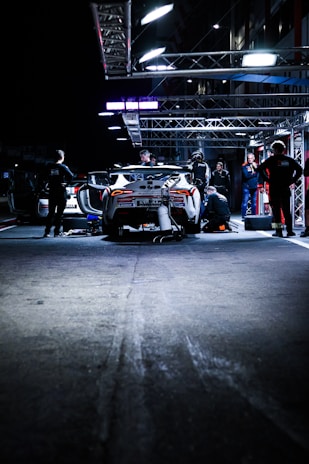 A race car is parked in a well-lit pit stop area at night. Several mechanics are working on the car, with some crouching near the wheels and others standing around. Various tools and equipment are scattered on the ground. The scene is illuminated by overhead lights, providing a contrast to the dark surroundings.